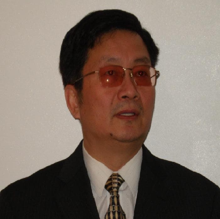East Asian man wearing orange tinted glasses, a dark suit, and a patterned tie.